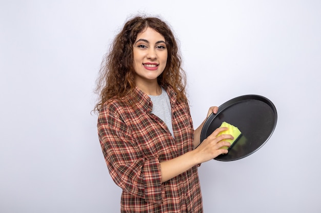 Sonriente joven mujer de la limpieza que se lava la bandeja con una esponja de limpieza aislado en la pared blanca