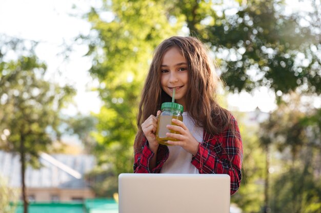 Sonriente joven mujer morena sentada en el parque con ordenador portátil