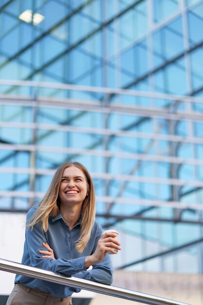 Sonriente joven mujer profesional tomando un café durante su jornada laboral completa. Ella sostiene un vaso de papel al aire libre cerca del edificio comercial mientras se relaja y disfruta de su bebida.