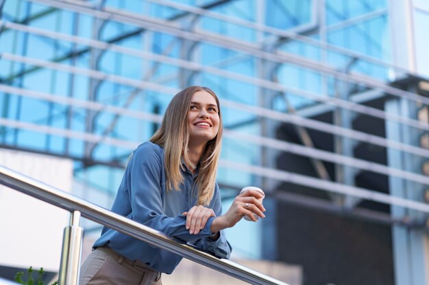 Sonriente joven mujer profesional tomando un café durante su jornada laboral completa. Ella sostiene un vaso de papel al aire libre cerca del edificio comercial mientras se relaja y disfruta de su bebida.