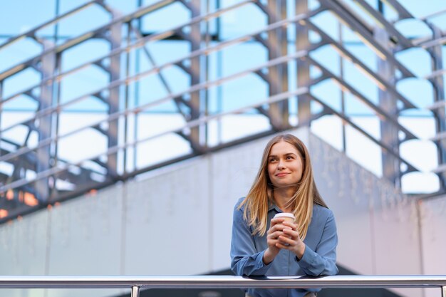 Sonriente joven mujer profesional tomando un café durante su jornada laboral completa. Ella sostiene un vaso de papel al aire libre cerca del edificio comercial mientras se relaja y disfruta de su bebida.