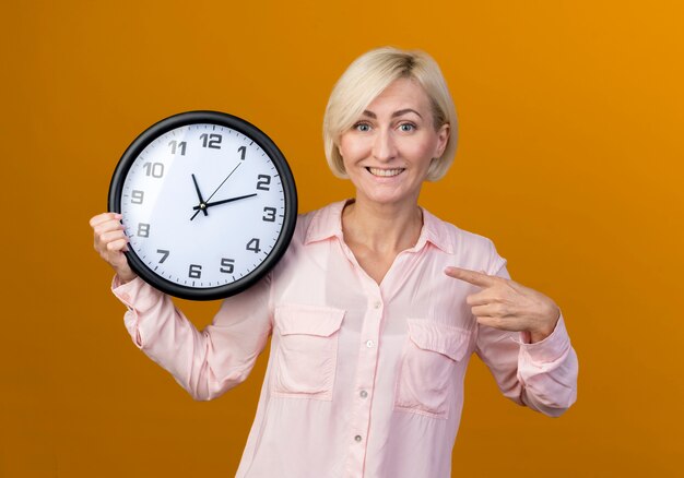 Sonriente joven mujer rubia eslava sosteniendo y apunta al reloj de pared aislado en naranja