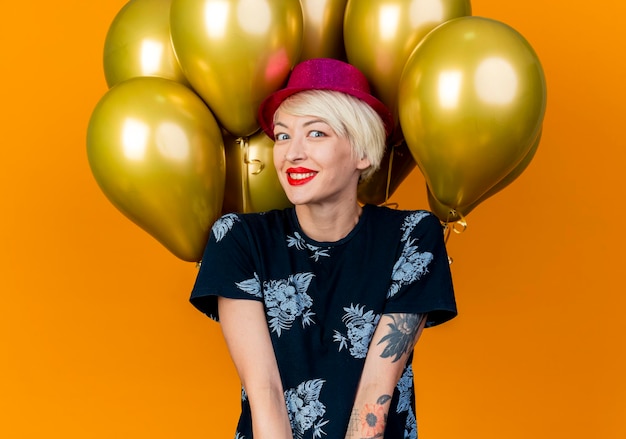 Foto gratuita sonriente joven mujer rubia fiesta con sombrero de fiesta de pie delante de globos mirando al frente aislado en la pared naranja