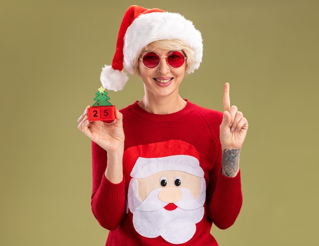Sonriente joven mujer rubia con sombrero de Navidad y suéter de Navidad de Papá Noel con gafas sosteniendo el juguete del árbol de Navidad con fecha mirando hacia arriba aislado en la pared verde oliva con espacio de copia