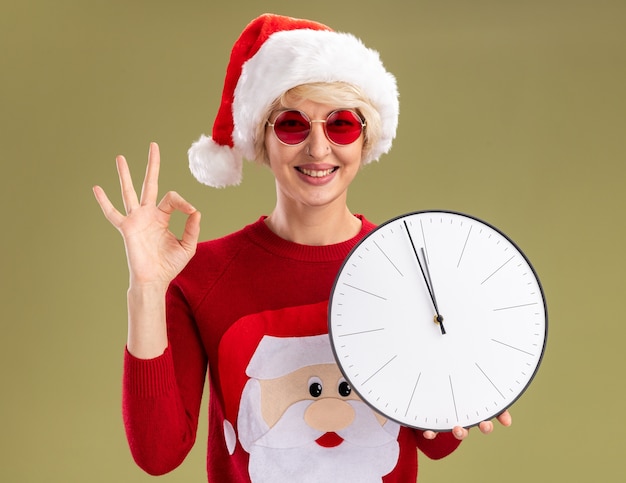 Foto gratuita sonriente joven mujer rubia con sombrero de navidad y suéter de navidad de papá noel con gafas sosteniendo reloj mirando haciendo bien firmar aislado en la pared verde oliva