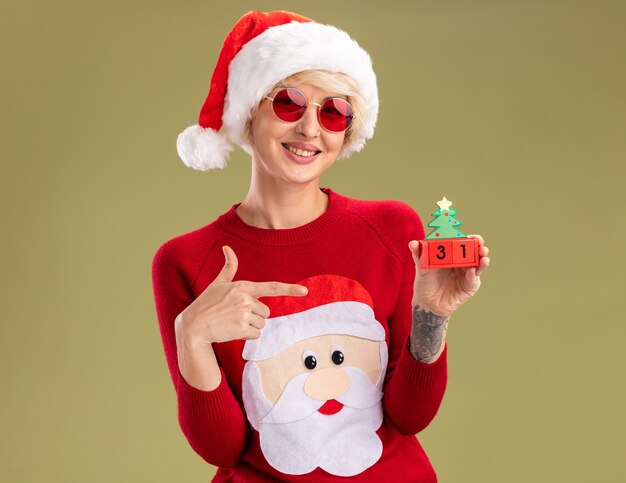 Sonriente joven mujer rubia con sombrero de navidad y suéter de navidad de santa claus con gafas sosteniendo y apuntando al árbol de navidad juguete con fecha mirando aislado en la pared verde oliva