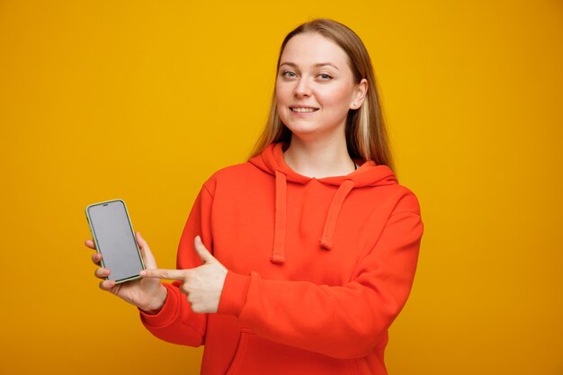 Sonriente joven mujer rubia sosteniendo y apuntando al teléfono móvil