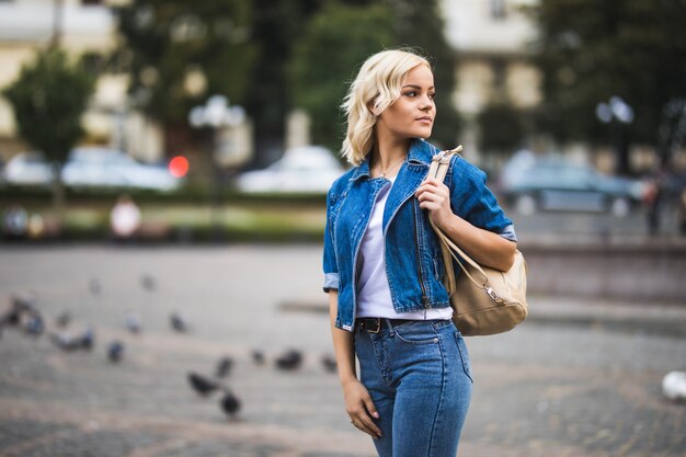 Sonriente joven mujer rubia en streetwalk square fontain vestida con blue jeans suite con bolsa en el hombro en un día soleado