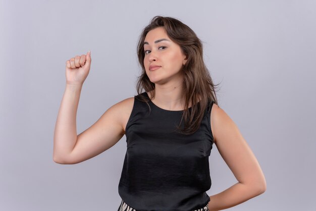 Sonriente joven mujer vestida con camiseta negra levantó el puño con una mano y el otro juego de palabras en la espalda en la pared blanca