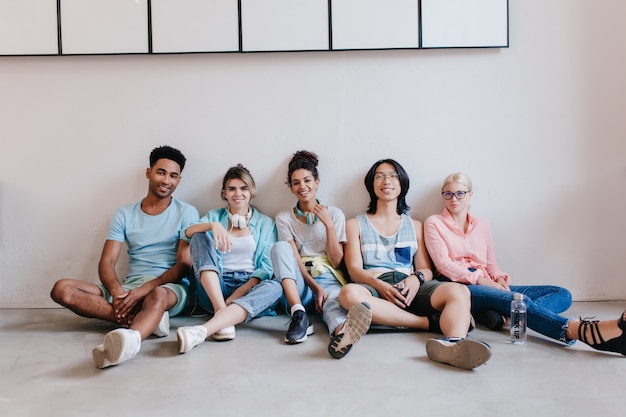 Foto gratuita sonriente joven negro con camisa azul sentado en el suelo junto a sus amigos. retrato interior de estudiantes internacionales esperando exámenes en el suelo.