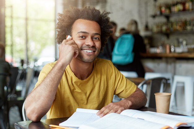 Sonriente joven negro sentado en la cafetería hablando por teléfono inteligente con amplia sonrisa y buen humor mientras descansa