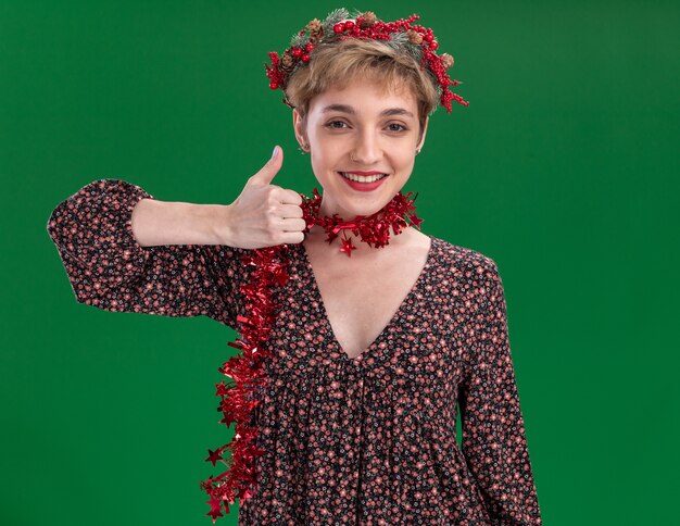 Sonriente joven niña bonita con corona de Navidad y guirnalda de oropel alrededor del cuello mostrando el pulgar hacia arriba aislado en la pared verde
