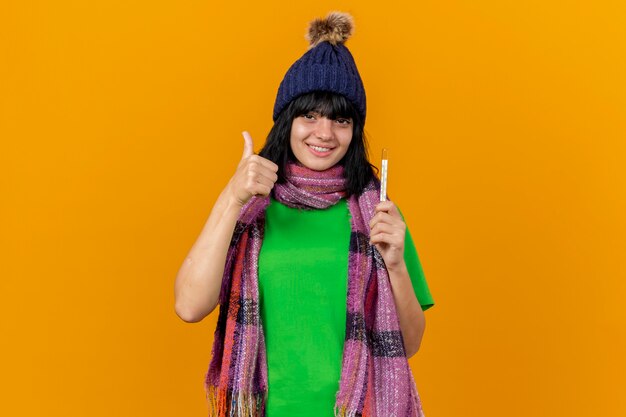 Sonriente joven niña caucásica enferma con gorro de invierno y bufanda sosteniendo el termómetro mostrando el pulgar hacia arriba aislado en la pared naranja con espacio de copia