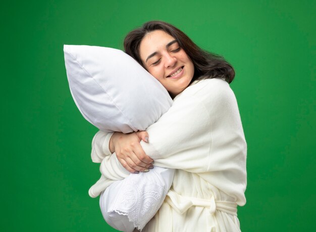 Sonriente joven niña enferma caucásica vistiendo bata de pie en la vista de perfil abrazando la almohada con los ojos cerrados aislado sobre fondo verde