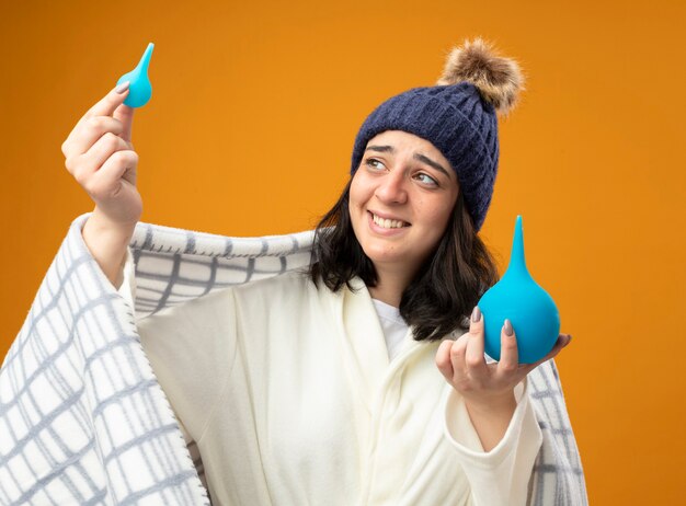 Sonriente joven niña enferma caucásica vistiendo bata sombrero de invierno envuelto en cuadros sosteniendo enemas levantando y mirando al pequeño aislado sobre fondo naranja