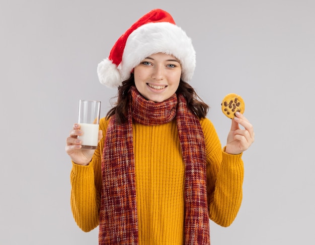 Sonriente joven niña eslava con gorro de Papá Noel y bufanda alrededor del cuello sostiene un vaso de leche y galletas aislado sobre fondo blanco con espacio de copia