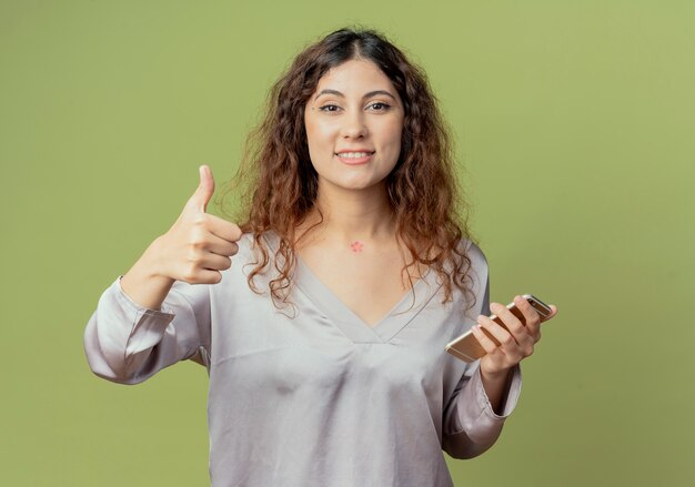 Sonriente joven oficinista bastante femenina sosteniendo el teléfono con el pulgar hacia arriba aislado en la pared verde oliva