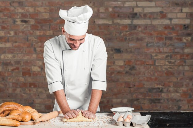 Sonriente joven panadero masculino amasar la masa en el mostrador de la cocina