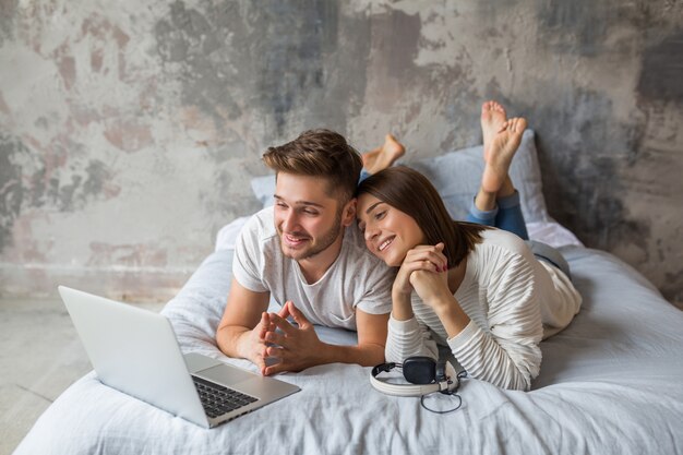 Sonriente joven pareja acostada en la cama en casa en ropa casual, mirando en la computadora portátil, hombre y mujer pasar tiempo feliz juntos, relajarse