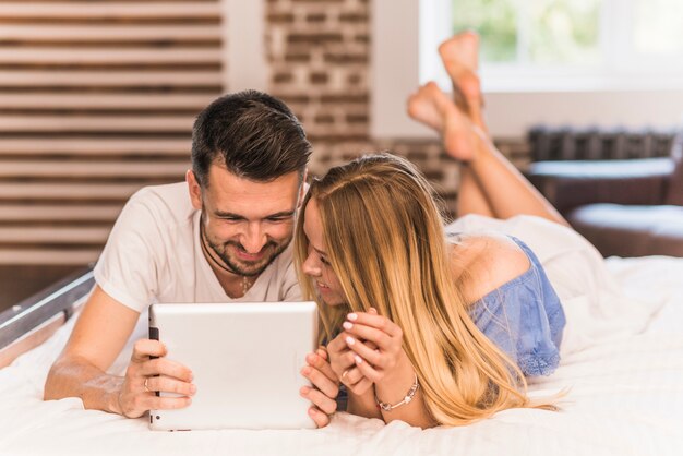 Sonriente joven pareja acostada en la cama mirando tableta digital