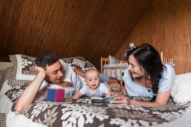 Sonriente joven pareja acostada en la cama mostrando libros a su bebé