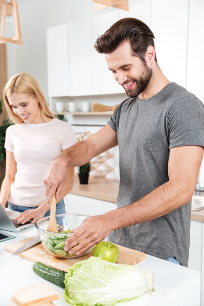 Sonriente joven pareja amorosa cocinar juntos usando laptop