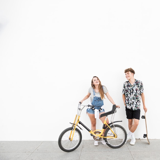 Sonriente joven pareja con bicicleta y patín contra la pared