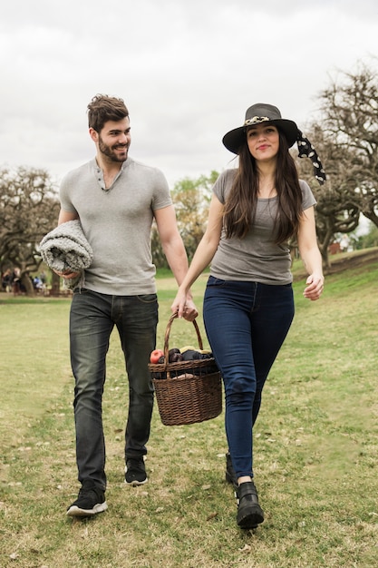 Sonriente joven pareja caminando con cesta de picnic en el parque