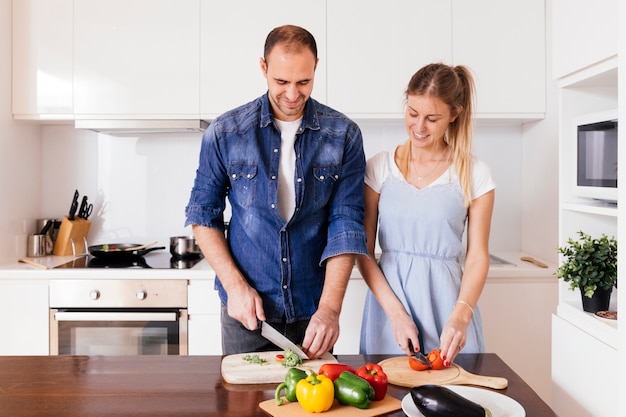 Sonriente joven pareja cortando las verduras con un cuchillo afilado en la cocina