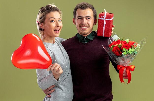 Sonriente joven pareja en el día de San Valentín se abrazaron sosteniendo un globo de corazón con regalos aislados sobre fondo verde oliva