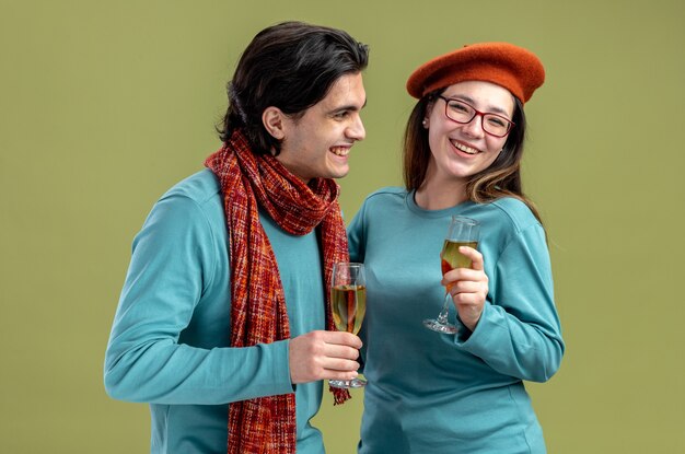 Sonriente joven pareja en el día de San Valentín chico con bufanda chica con sombrero sosteniendo una copa de champán aislado sobre fondo verde oliva