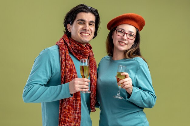 Sonriente joven pareja en el día de San Valentín chico con bufanda chica con sombrero sosteniendo una copa de champán aislado sobre fondo verde oliva