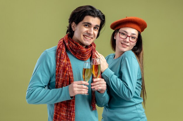 Sonriente joven pareja en el día de San Valentín chico con bufanda chica con sombrero sosteniendo una copa de champán aislado sobre fondo verde oliva