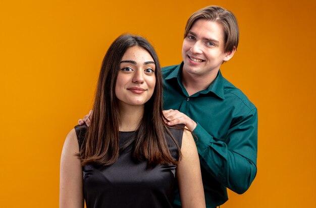 Sonriente joven pareja en el día de San Valentín chico de pie detrás de la chica poniendo la mano sobre el hombro aislado sobre fondo naranja