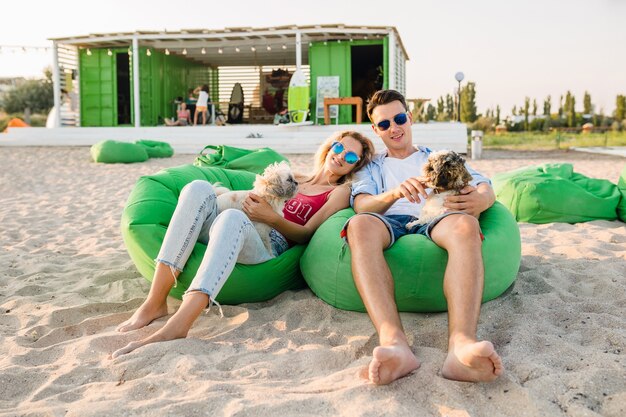 Sonriente joven pareja divirtiéndose en la playa jugando con perros de raza shih-tsu, sentado en una bolsa de frijoles verdes
