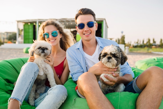 Sonriente joven pareja divirtiéndose en la playa jugando con perros de raza shih-tsu, sentado en una bolsa de frijoles verdes