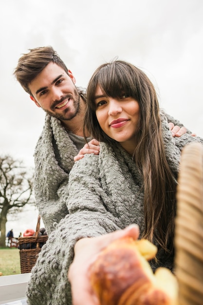 Sonriente joven pareja envuelta en una manta gris dando croissants al horno