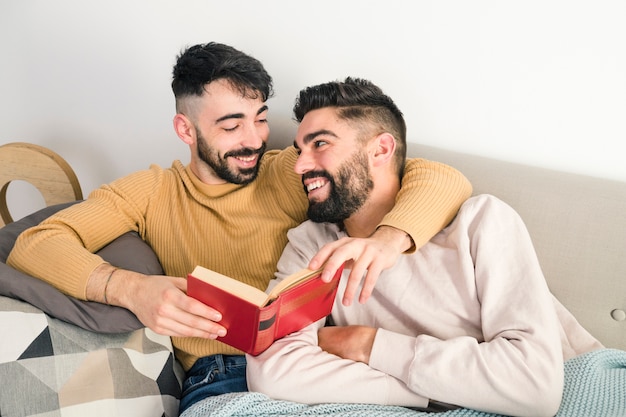 Sonriente joven pareja gay mirando el uno al otro mientras lee el libro