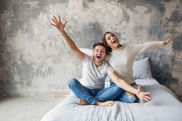 Sonriente joven pareja sentada en la cama en casa en ropa casual, hombre y mujer divirtiéndose juntos, loca emoción positiva, feliz, levantando las manos