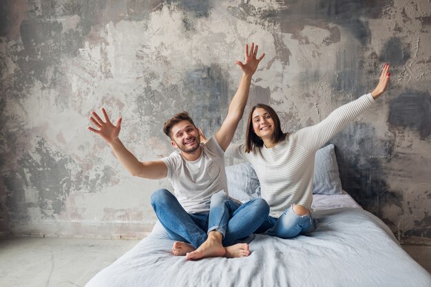 Sonriente joven pareja sentada en la cama en casa en ropa casual, hombre y mujer divirtiéndose juntos, loca emoción positiva, feliz, levantando las manos