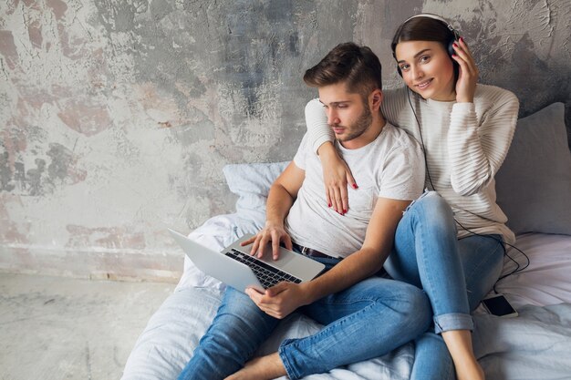 Sonriente joven pareja sentada en la cama en casa en ropa casual, hombre trabajando independiente en la computadora portátil, mujer escuchando música en auriculares, pasando tiempo feliz juntos