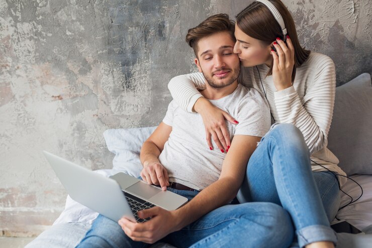 Foto gratuito sonriente joven pareja sentada en la cama en casa en ropa casual, hombre trabajando independiente en la computadora portátil, mujer escuchando música en auriculares, pasando tiempo feliz juntos