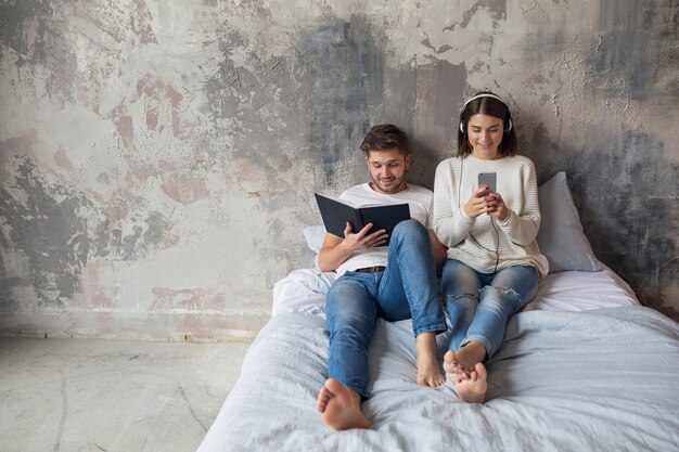 Sonriente joven pareja sentada en la cama en casa en ropa casual leyendo libro con jeans, libro de lectura de hombre, mujer escuchando música en auriculares, pasando tiempo romántico juntos