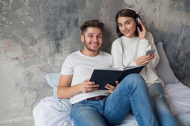 Sonriente joven pareja sentada en la cama en casa en ropa casual leyendo libro con jeans, libro de lectura de hombre, mujer escuchando música en auriculares, pasando tiempo romántico juntos