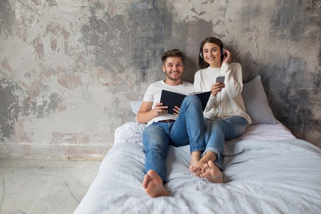 Sonriente joven pareja sentada en la cama en casa en ropa casual leyendo libro con jeans, libro de lectura de hombre, mujer escuchando música en auriculares, pasando tiempo romántico juntos