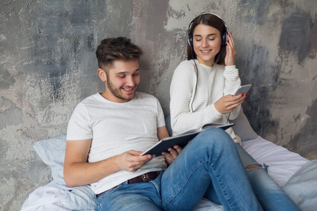 Sonriente joven pareja sentada en la cama en casa en ropa casual leyendo libro con jeans, libro de lectura de hombre, mujer escuchando música en auriculares, pasando tiempo romántico juntos