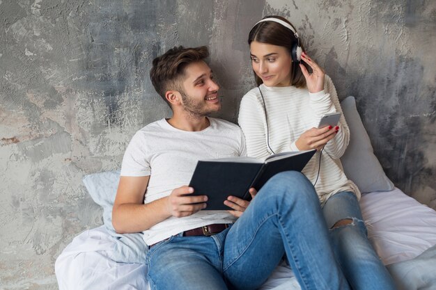 Sonriente joven pareja sentada en la cama en casa en ropa casual leyendo libro con jeans, libro de lectura de hombre, mujer escuchando música en auriculares, pasando tiempo romántico juntos