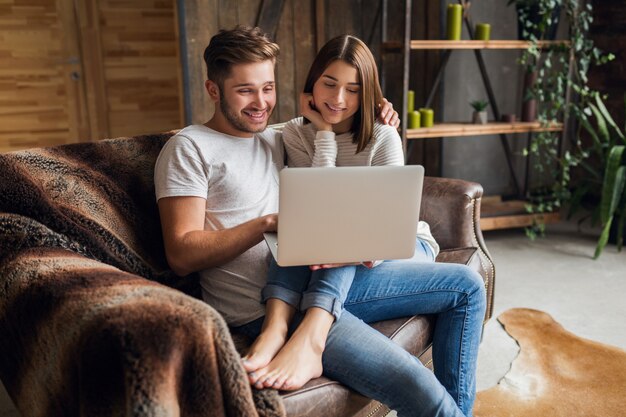 Sonriente joven pareja sentada en el sofá en casa en ropa casual, amor y romance, mujer y hombre abrazados, vistiendo jeans, pasando tiempo de relax juntos, sosteniendo la computadora portátil