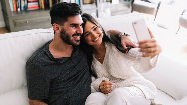 Sonriente joven pareja sentada en el sofá tomando selfie