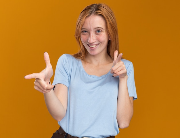 Sonriente joven pelirroja pelirroja con pecas apuntando a la cámara con las dos manos aisladas en la pared naranja con espacio de copia